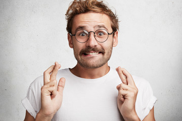 Positive bearded young male wears round spectacles, crosses fingers and bites lips has great hope...