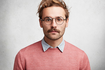 Isolated shot of confident bearded male wears round spectacles and sweater, looks confidently into camera, listens to important information, poses against white concrete background. Facial expressions