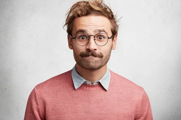 Fotobehang Clumsy male geek presses lips, looks in bewilderment, feels embarrassed in presence of beautiful woman, wears round big spectacles, isolated over white concrete background. Excited emotional male © Wayhome Studio