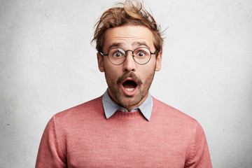 Indoor shot of amazed shocked young man with mustache, stares through round spectacles, being surprised to hear unexcpected news, can`t believe eyes, isolated over white concrete background.