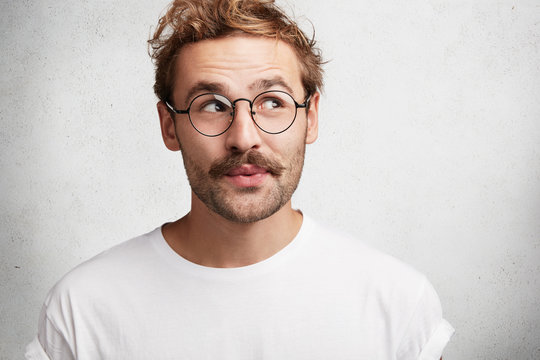 Photo of pleased dreamy male wonk with trendy hairdo, wears spectacles and casual white t shirt, imagines something pleasant, looks upwards with intriguing expression, isolated on concrete wall
