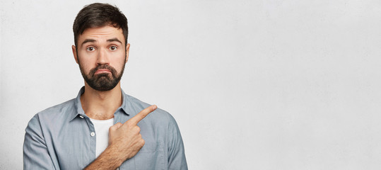 Horizontal portrait of bearded male model looks with bewilderment, indicates at blank copy space for text as notices something strange. Pleasant looking unshaven guy advertizes new product indoor