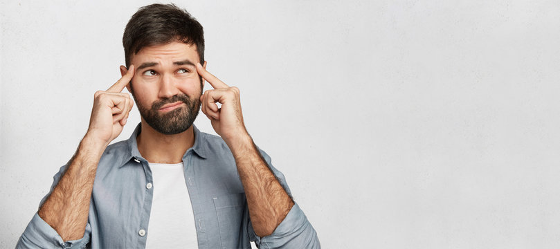 Pleasant looking bearded young male tries to find solution in mind, keeps figers on temples for concentration, looks aside, poses against white concrete wall with copy space for promotional text