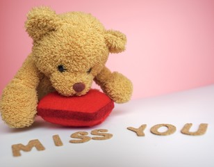 Close up of cute brown teddy bear leaning on bright red heart looking at words miss you on white table top with pink background. Emotion concept. 