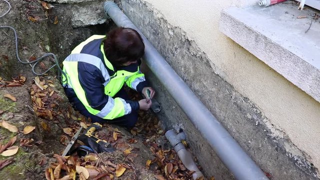 A female plumber makes measuring water pipes for replacement outside the house.