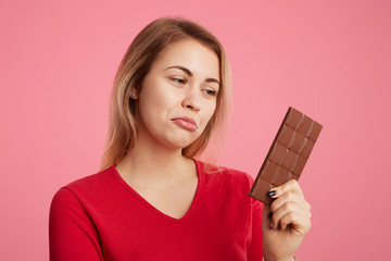 Attractive young woman looks with discontent expression at sweet bar of chocolate, keeps to diet, can`t eat it to be slim and sporty, curves lower lip, isolated over pink background. Eating sweets