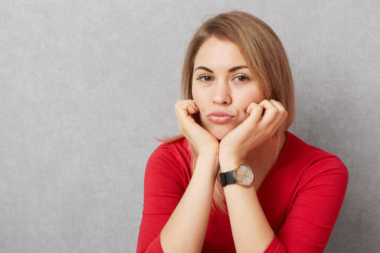 Photo of sad pretty woman leans at hands, pouts lips, wears fashionable watch and red sweater, looks with upset expression into camera, isolated over grey background. Facial expressions concept