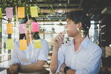 man & woman discussing creative idea with adhesive notes on glass wall at workplace. Sticky note paper reminder schedule at office. business, brainstorming, creativity concept