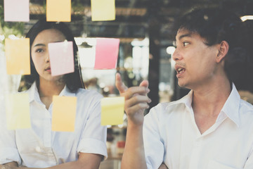 man & woman discussing creative idea with adhesive notes on glass wall at workplace. Sticky note paper reminder schedule at office. business, brainstorming, creativity concept