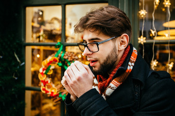 Outdoor portrait of beard man in winter Christmas background and warmed the hands of breath. Guy posing on street. Travel mood concept.