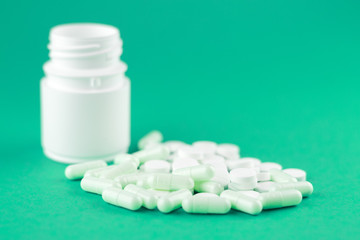 Close up white pill bottle with spilled out pills and capsules on emerald green background with copy space. Focus on foreground, soft bokeh. Pharmacy drugstore concept