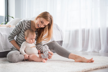 Full length of smiling mother sitting on soft carpet and showing teddy bear to his lovely daughter