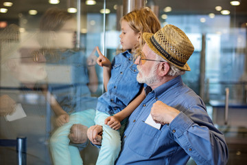 Side view profile of concentrated grandfather is standing at terminal hall with his grandoughter on hands. He is putting tickets at pocket of his shirt while girl touching glass wall. Selective focus