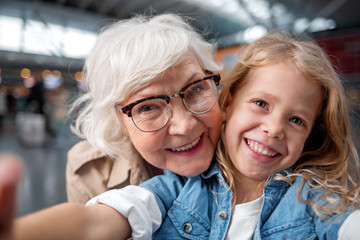 Cool picture. Close-up portrait of joyful happy elderly grandmother is taking selfie with her adorable granddaughter. They are looking at camera with wide smile while standing at airport hall