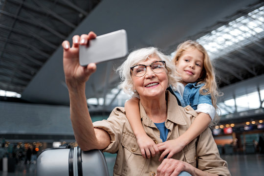 Nice photo. Portrait of cheerful aged charming grandmother is standing at airport lounge with her little granddaughter and looking at screen of mobile phone while taking selfie
