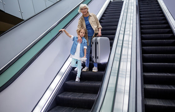 Low Angle Full Length Portrait Of Cheerful Elegant Senior Grandmother Is Standing On Escalator At International Airport With Her Granddaughter While Hugging Her And Holding Suitcase. Copy Space