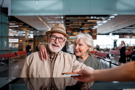 Have nice trip. Portrait of cheerful gray-haired couple is standing at international airport near registration desk. Old man is taking his tickets while aged woman is looking at him with love