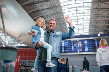 Our joyful trip. Low angle portrait of cheerful middle-aged father is holding his little daughter while standing at international airport and waving. They are looking at camera with wide smile