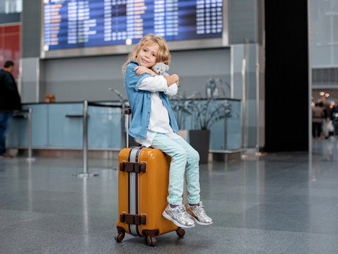Love my bear. Portrait of adorable little child is sitting on orange suitcase at airport lounge and hugging her lovely toy. She is looking at camera with joy. Electronic timetable in backgraund