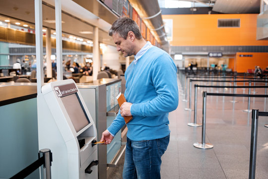 Side view profile of serious mature man is standing near self-service check-in kiosk at international airport building. He is using his credit card to making payment for flight tickets