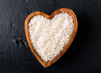 Isolated handful of raw white rice in the wood bowl in the shape of heart on black stone background