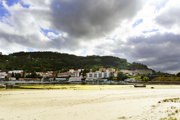 Fototapeta na wymiar Beach and town of Pontedeume in La Coruna, Spain on the other side of its bay. Sky with clouds threatening rain and lots of vegetation next to the town. A small boat stranded in the sand