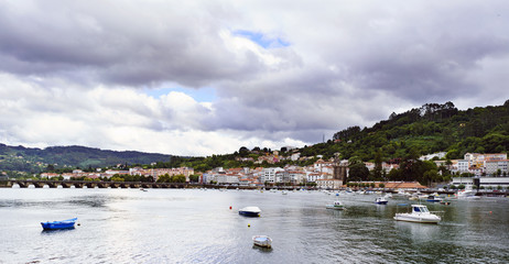 Fototapeta na wymiar View of the Eume estuary with the stone bridge that crosses it to the bottom and anchored pleasure boats. The village of Pontedeume, Galicia, Spain in the background and a sky covered with clouds