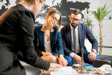 Young businesscouple choosing a trip with agent sitting at the travel agency office with world map...