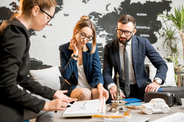 Young businesscouple choosing a trip with agent sitting at the travel agency office with world map...
