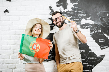 Couple of young travelers standing with portuguese flag near the wall with world map, dreaming about summer vacations in Portugal