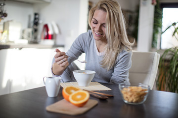 Obraz na płótnie Canvas Beautiful blond caucasian woman posing in her kitchen, while drinking coffee or tea and eating a healthy breakfast meal full of cereal and other healthy foods, including fruit