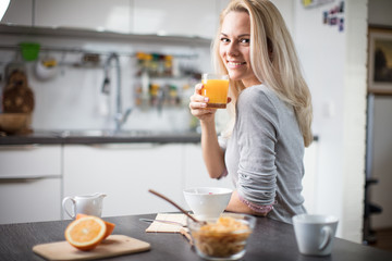 Beautiful blond  caucasian woman posing in her kitchen, while drinking coffee or tea and eating a healthy breakfast meal full of cereal and other healthy foods, including fruit