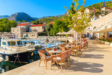Fototapeta premium BOL PORT, BRAC ISLAND - SEP 14, 2017: restaurant tables in Bol port with fishing boats on late summer afternoon, Brac island, Croatia.