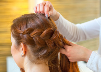 Naklejka premium Beautiful girl with long red hair, hairdresser weaves a braid, in a beauty salon. Professional hair care and creating hairstyles.