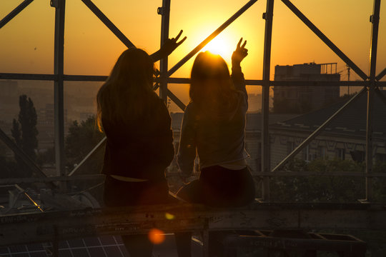 Two girl silhouettes with V victory ,OK, super  gesture  sitting on the top of a high building, with a sun flare , sunrise  in front of them . Concept of best friend forever girl teen .
