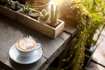 White cup of hot coffee with the white cream and white plate on the wooden table in the restaurant or coffee shop with blurred garden background, cappuccino