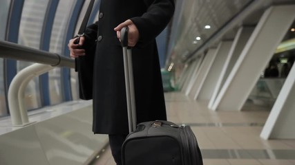Stylish male brunette in black coat is holding suitcase handle in airport. Passenger in strict clothing after flight announcement goes to exit and pulls luggage.