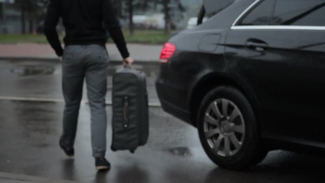 Man is loading large suitcase with handle in trunk of car. In rainy weather driver of car removes luggage from person in coat in transport section.