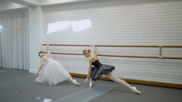 Female ballerinas in tutu take different dance positions at machine. Two woman dancers in black and white dress are training in a spacious ballet studio.