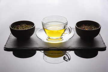 Minimalist tea set up.  Cup of tea and dried organic tea herbs in pots, on top of the reflective table, against beautiful day light.  