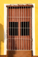 Brown shuttered window behind white metal bars on yellow and white wall