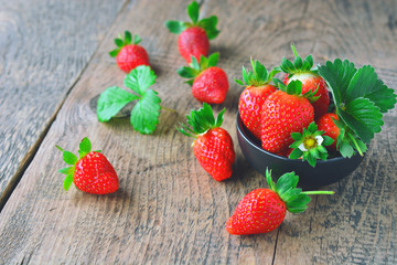 Fresh red strawberries in a black bowl and leaf on wooden table in the background.
