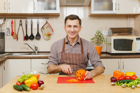 Handsome smiling caucasian young man in an apron, brown shirt sitting at table, cuts vegetable for salad with knife in light kitchen. Dieting concept. Healthy lifestyle. Cooking at home. Prepare food.