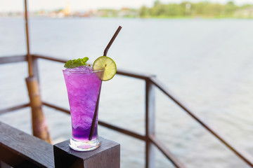 Butterfly pea iced soft drinks with lemon slice and peppermint on top and on the wooden balcony at the riverside restaurant.Thailand.