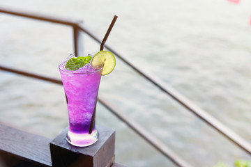 Butterfly pea iced soft drinks with lemon slice and peppermint on top and on the wooden balcony at the riverside restaurant.Thailand.