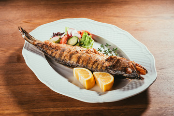 Baked fish with lemon and herbs on white plate with salad on wooden table