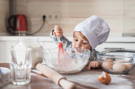 happy little kid in chef hat mixturing dough with whisk at kitchen