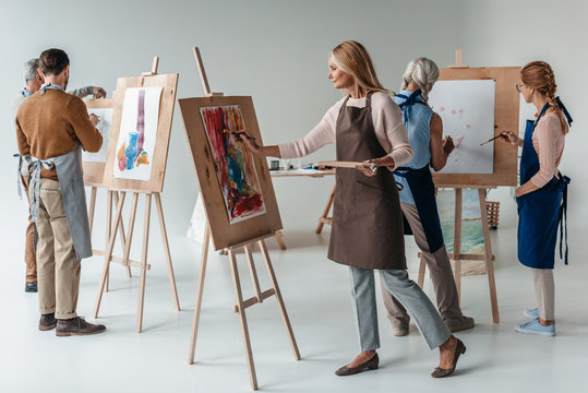 group of male and female adult students in aprons painting together on easels in art class