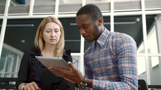 White woman and African American man look closely at tablet on hands. Adult lady and young guy are seriously studying monitor screen on wireless device.