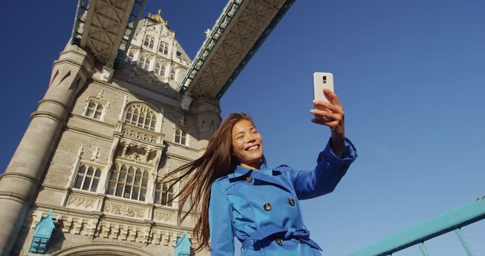 England woman taking selfie though mobile smart phone on London Tower Bridge. Tourist is smiling while taking smartphone selfportrait. visiting famous places in UK. Young professional in trench coat.